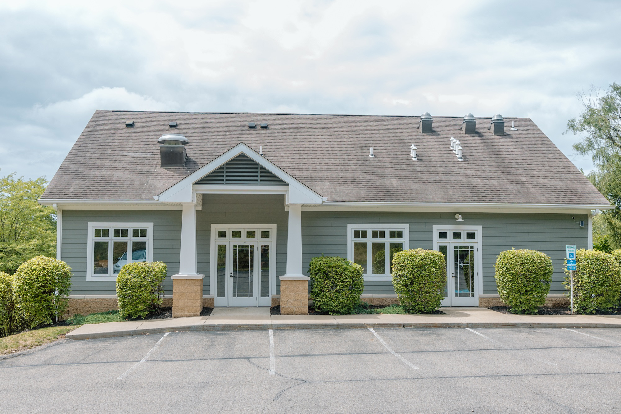 The image shows a single-story building with a prominent front entrance, featuring a white facade and a covered porch. The structure has a peaked roof with dark shingles and is topped with a small cupola. There are trees visible in the background, and the sky appears overcast.