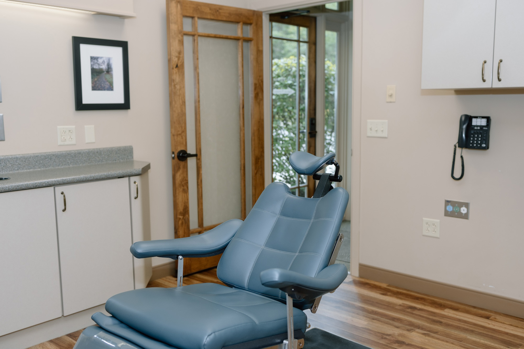 The image shows a modern dental office interior with a blue reclining dental chair, a counter with a sink and cupboards, a wall-mounted television monitor, and a door leading to another room.