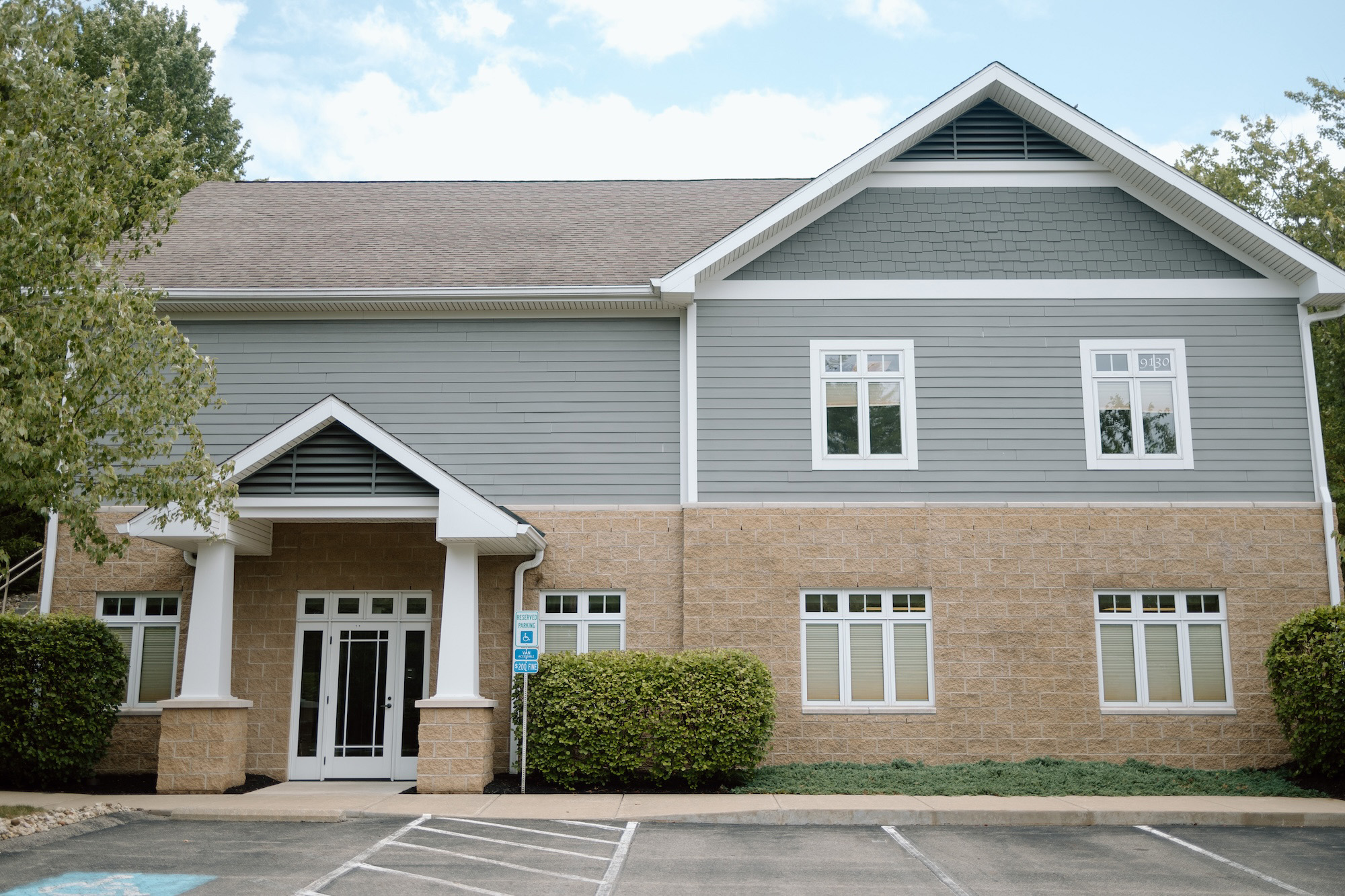 The image depicts a two-story building with a white exterior, featuring a gray roof and a covered entrance with columns. A sign is visible on the front of the building, but the text is not legible in the image.