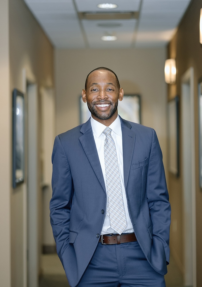 A man in a suit stands confidently in an office environment, smiling towards the camera.