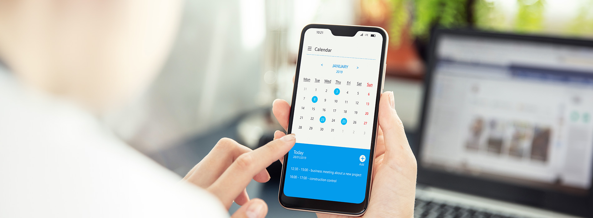 This is a color photograph showing a person holding a smartphone with a user interface displaying a calendar and a weather forecast. The screen shows an app with the time set to 10 45, and the background includes a blurred image of a laptop and a desk with various items.