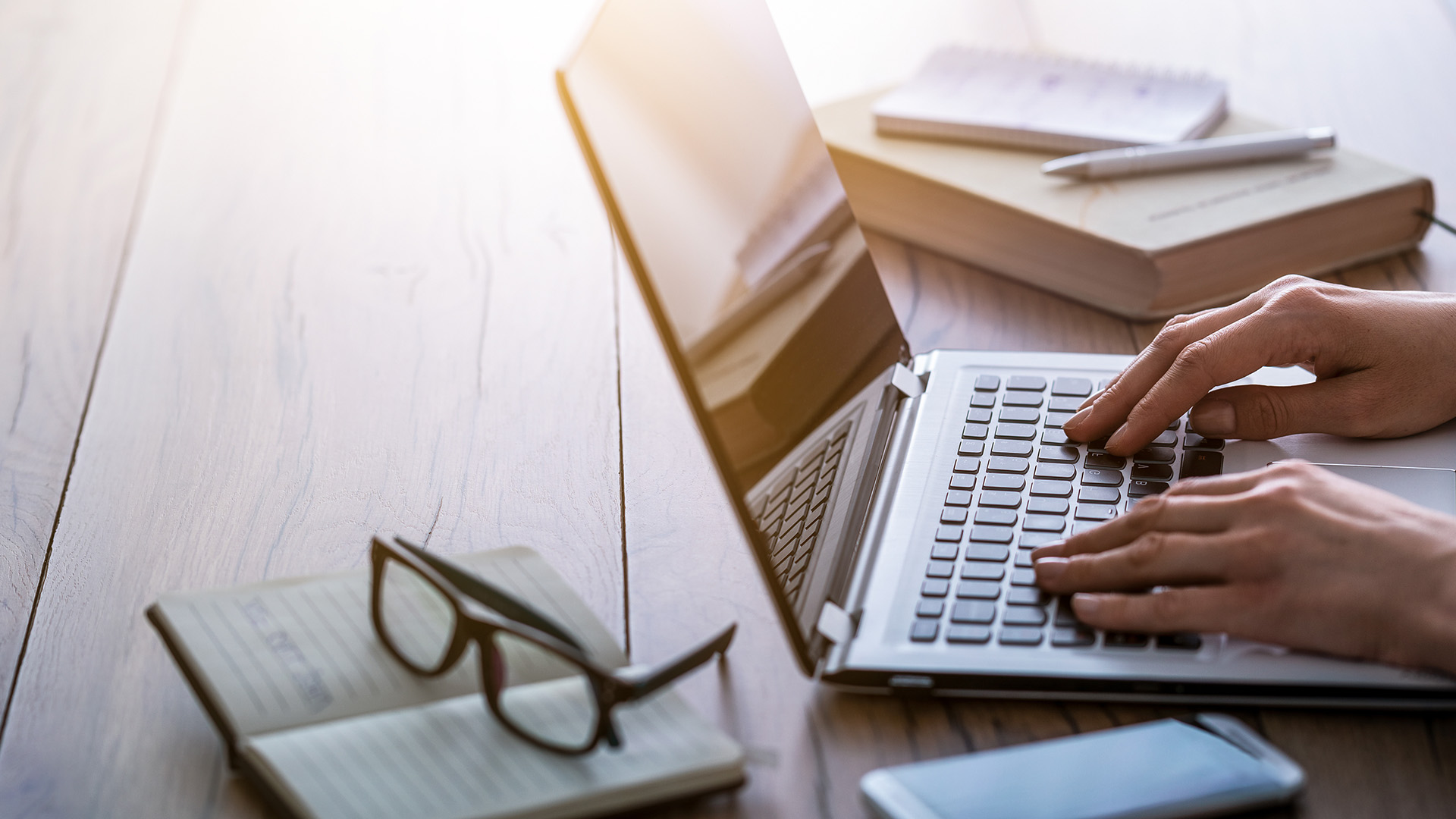 A person using a laptop at a desk with books and papers, set against a blurred background of a sunlit room.