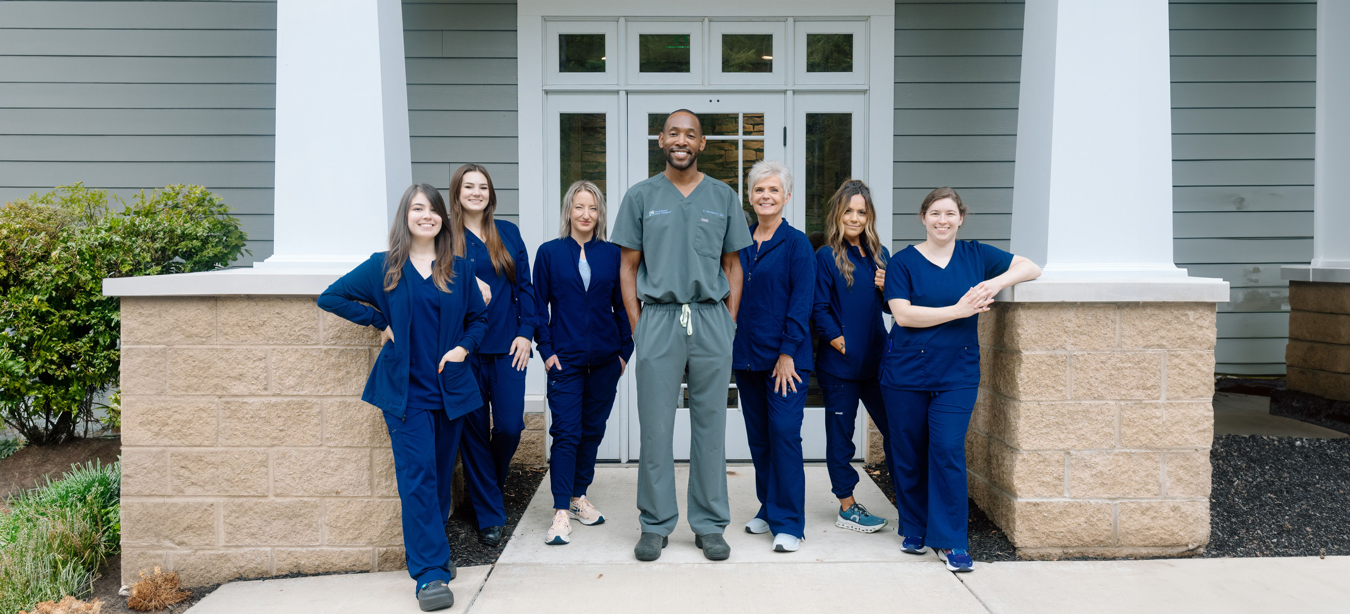 A group of five people, likely nurses or healthcare workers, standing in front of a building with a white pillar and a brick wall.