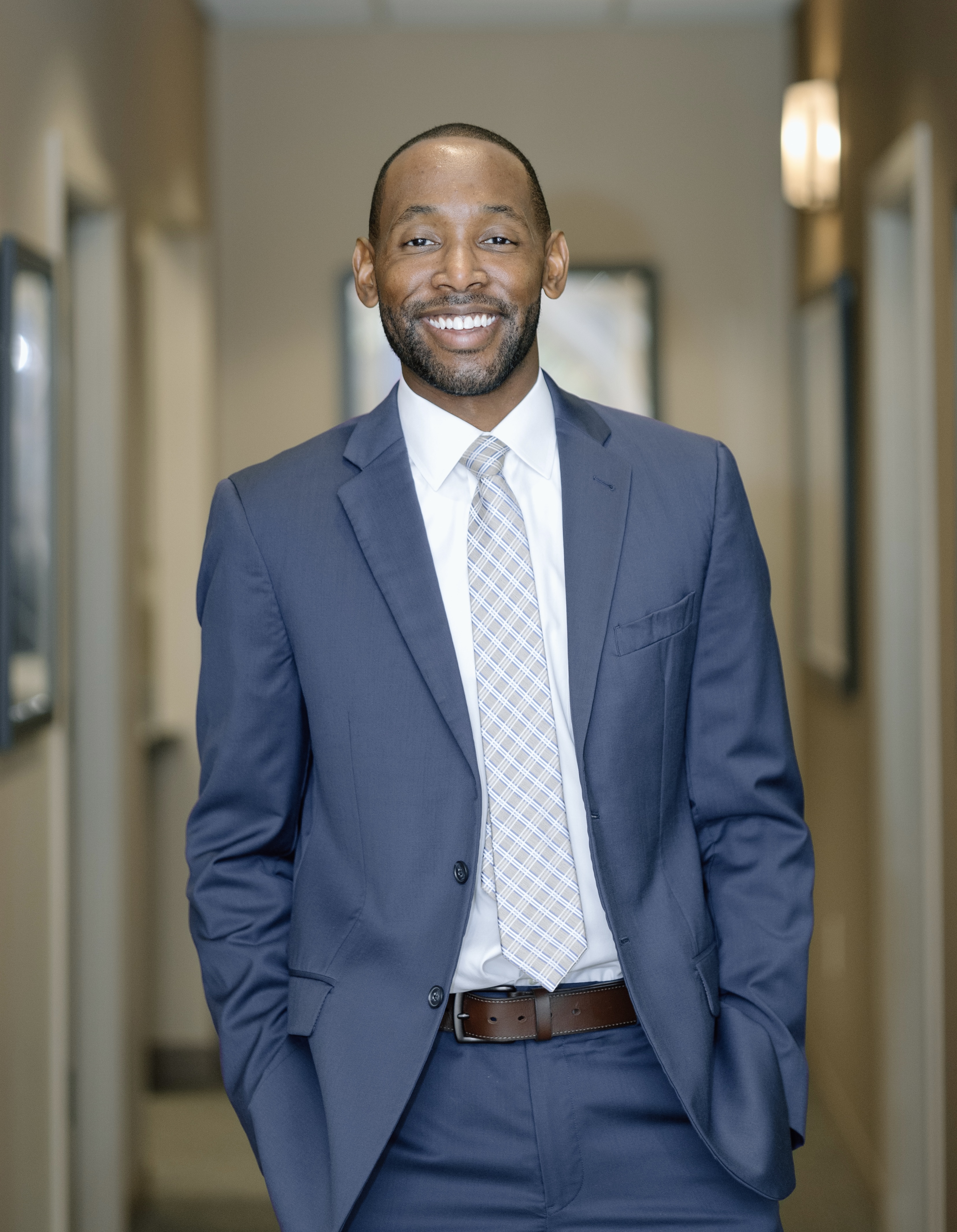 The image features a man in a suit standing inside an office, smiling directly at the camera.