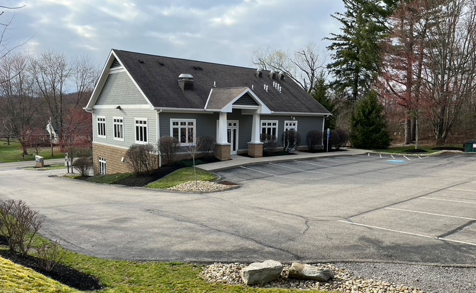 A large, single-story building with a gray roof and a prominent front entrance, featuring a covered porch with a bench. The structure is situated in a parking lot with multiple spaces, adjacent to a wooded area with trees and a pathway.