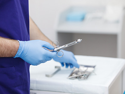 A medical professional wearing protective gloves and holding a pair of surgical instruments, standing in front of a sterile tray with additional instruments.