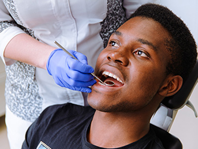 A man in a dental chair receiving dental treatment, with a dental hygienist standing behind him, assisting with the procedure.