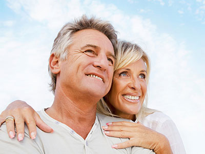 The image shows a man and a woman embracing each other, with the man facing the camera and both individuals smiling. They appear to be older adults enjoying a moment together outdoors during the daytime.