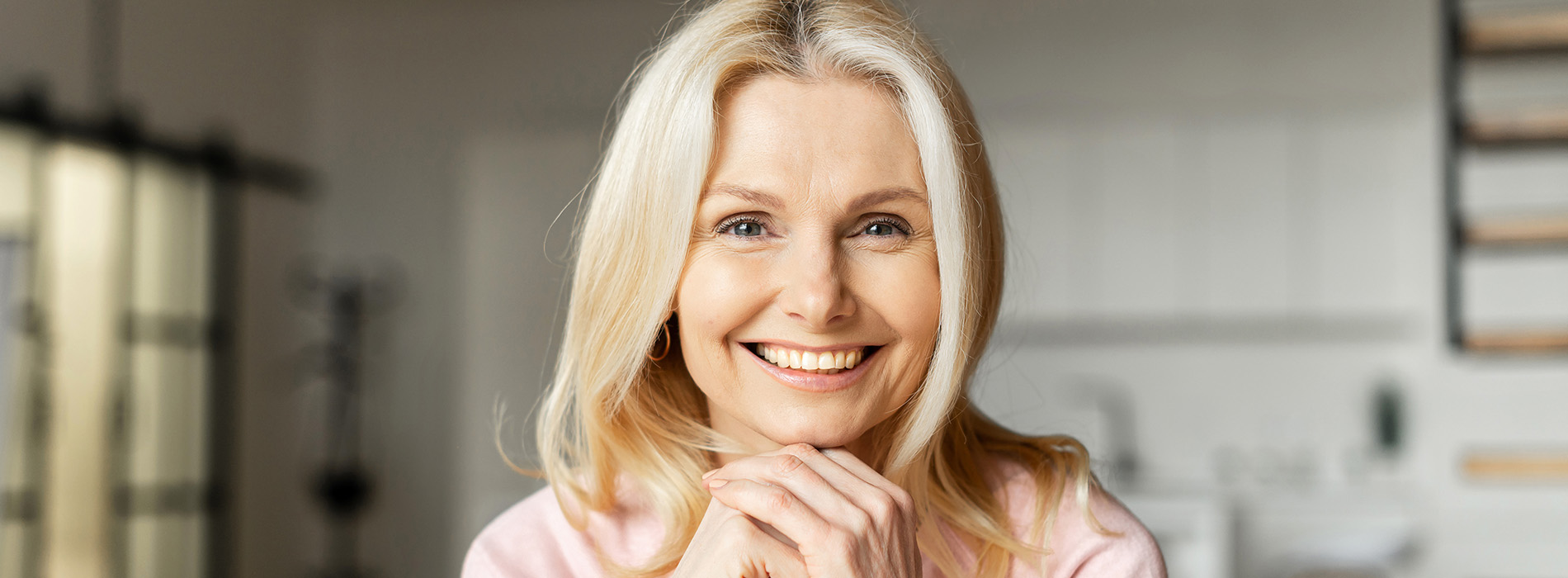 A smiling woman with blonde hair, wearing a pink top, stands in a kitchen with white cabinets and appliances behind her.