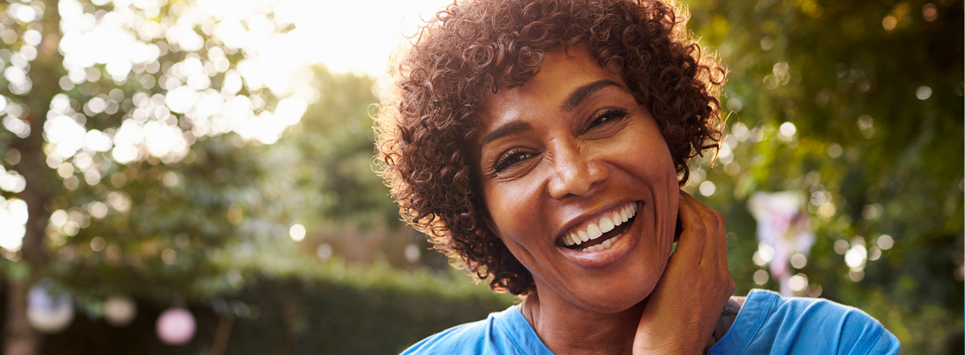 A smiling woman with curly hair, wearing a blue top, stands outdoors during daylight.