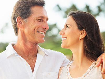 The image shows a man and woman, likely in their middle to older years, smiling and embracing each other outdoors during the day.