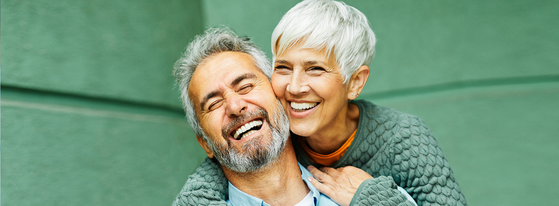 A man and a woman are smiling, with the man hugging the woman from behind. They appear to be an older couple sharing a joyful moment together.