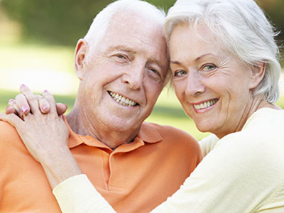 An elderly couple embracing each other outdoors, with the man wearing an orange shirt and both smiling.