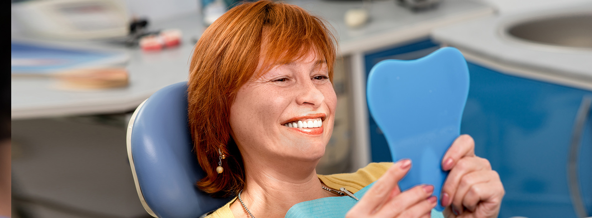 A smiling woman with red hair, wearing a yellow top, is seated in a dental chair holding a blue object, possibly an impression tray or a dental tool.