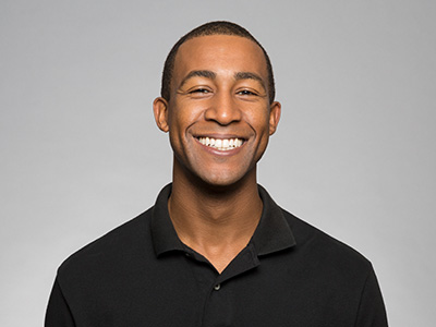 The image shows a smiling man wearing a dark shirt and standing against a white background. He appears to be middle-aged with short hair and is looking directly at the camera.