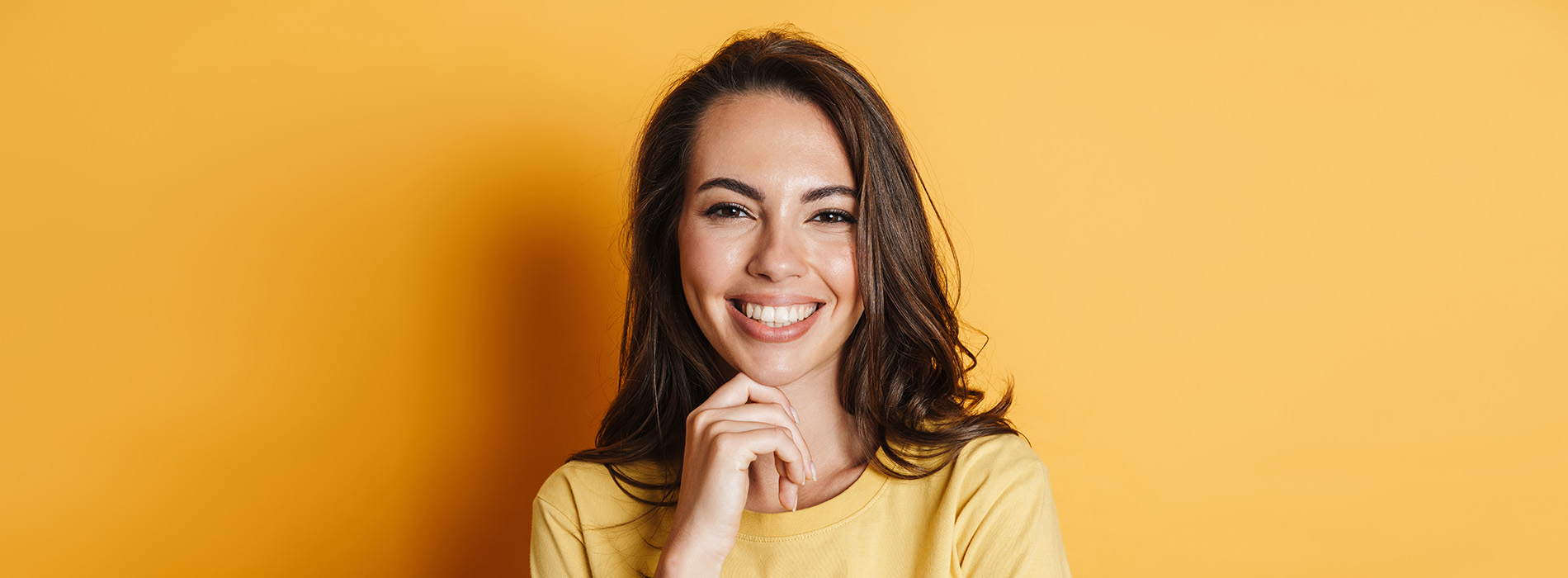 The image shows a smiling woman with dark hair, wearing a yellow top, standing against a bright yellow background.